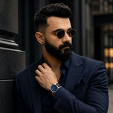 A man with a beard and sunglasses wearing a navy blue suit and matching shirt, posing outdoors against a dark stone wall while wearing a navy watch.
