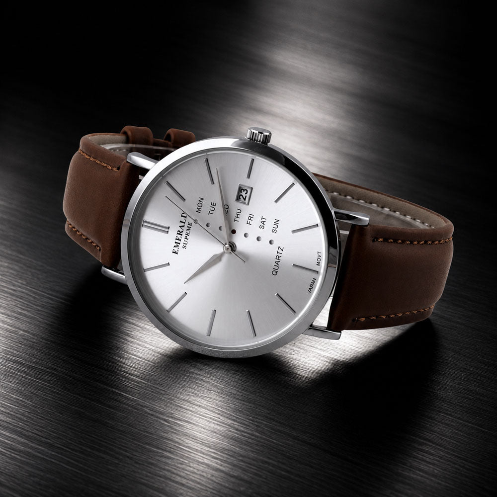 A luxury wrist watch with a silver-toned stainless steel case, a white dial featuring a day and date window, and a brown leather strap, displayed on a dark brushed metal surface.