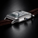 A close-up of a silver-tone rectangular watch with a brown leather strap, shown resting on a dark brushed surface. The watch has a clear crystal, Roman numeral hour markers, and a blue accent on the crown.
