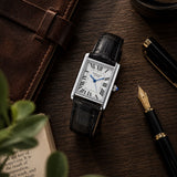 A Cartier Tank watch with a silver-toned rectangular case, white dial, black leather strap, and blue crown jewel, styled on a dark wood desk with a brown leather notebook, pen, and book.