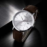 A wristwatch with a silver-tone case, white dial, and brown leather strap, photographed on a dark brushed-metal surface. The dial shows the brand name EMERALD and additional day/date text.