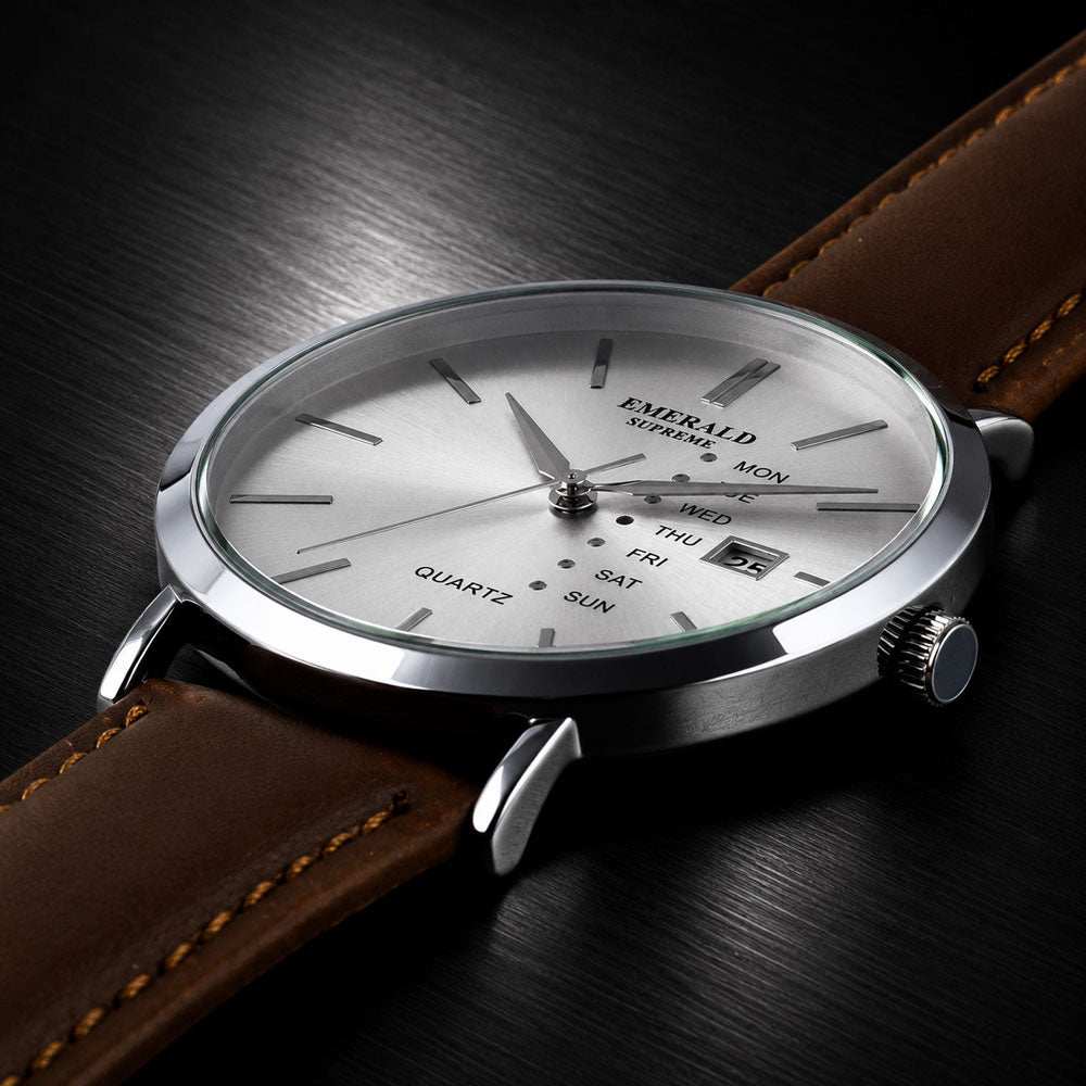 Emerald watch with a silver-toned stainless steel case, white dial, and brown leather strap, shown at an angle on a dark wood background.