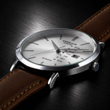 Emerald watch with a silver-toned stainless steel case, white dial, and brown leather strap, shown at an angle on a dark wood background.