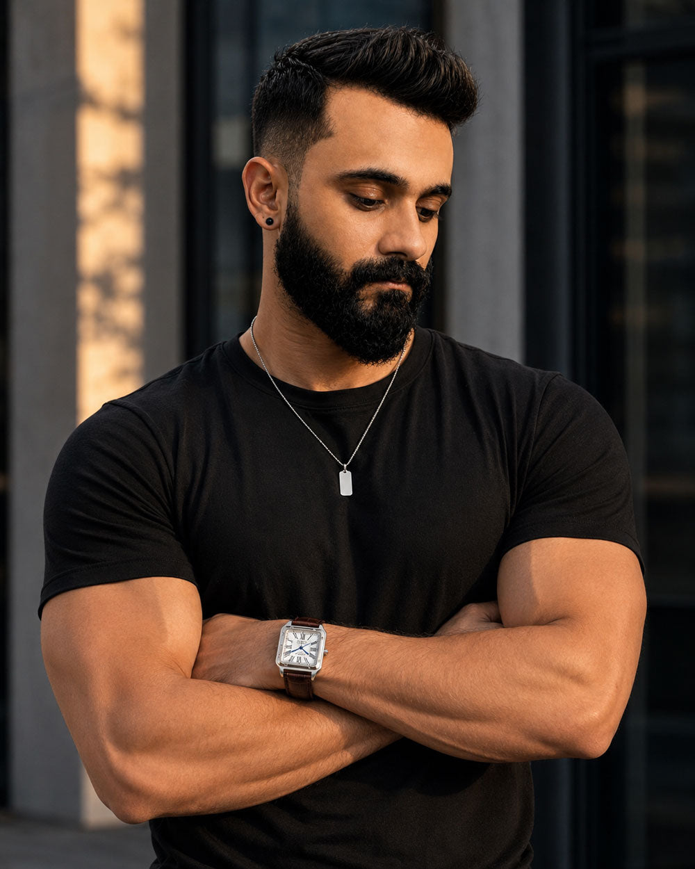 A muscular man with a beard and styled haircut wearing a black t-shirt, silver necklace, and a silver watch with a square face, standing with arms crossed.