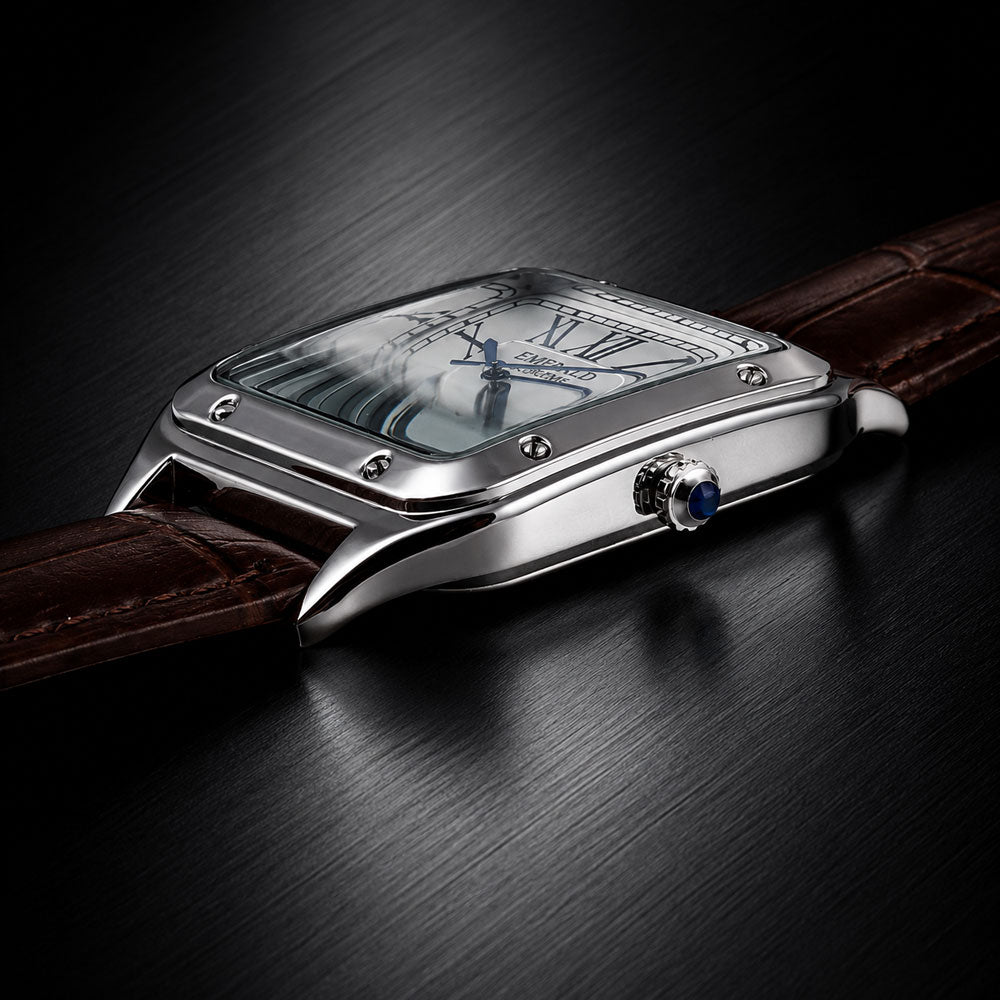 A close-up of a silver-tone rectangular watch with a brown leather strap, shown resting on a dark brushed surface. The watch has a clear crystal, Roman numeral hour markers, and a blue accent on the crown.